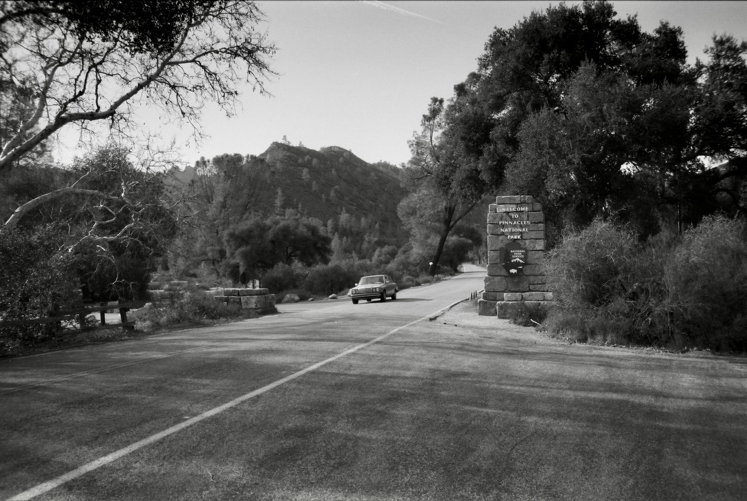 Black-and-white view of a road entering Pinnacles National Park, with a car approaching a stone entrance sign