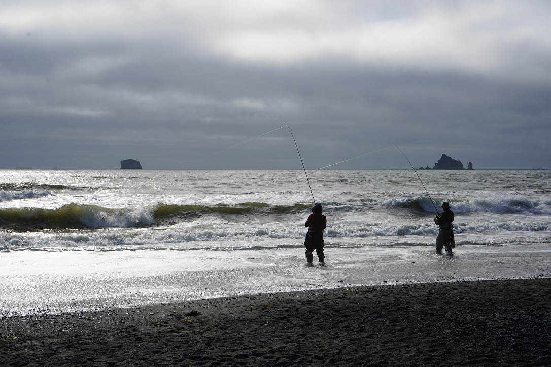 Two silhouetted anglers surf fishing in rough water beneath a gray sky, with sea stacks on the horizon
